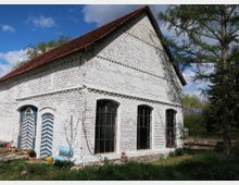 Ein weiß gestrichenes Backsteingebäude mit rotem Satteldach, großen rechteckigen Fenstern und zwei blau-weiß gestreiften Türen. Im Vordergrund sind ein kleiner Garten mit Blumen sowie ein Baum und andere Pflanzen zu sehen; im Hintergrund ist eine ländliche Umgebung mit weiteren Gebäuden und Solarmodulen erkennbar.