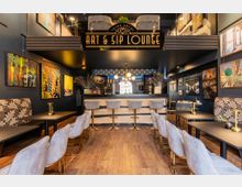 Modernes Café-Interieur mit Bar in der Mitte, dekoriert mit einer Mischung aus abstrakten und fotografischen Kunstwerken an den Wänden. Sitzgelegenheiten umfassen Polsterbänke mit floralen Mustern und gepolsterte Stühle an Holztischen, in einem Raum mit warmem Holzfußboden und einer dunklen Decke.