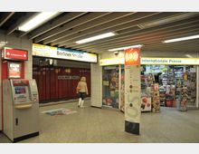 U-Bahnhof mit Schild "Berliner Straße", einer Notruf- und Informationssäule sowie einem Kiosk mit der Aufschrift "Internationale Presse". Links steht ein Geldautomat, und eine Person geht in Richtung der Bahnsteige.