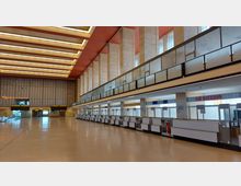 Große, offene Flughafenhalle im Retro-Stil mit hohen Decken, mehreren Säulen und Reihen verlassener Check-in-Schalter. An der Rückwand befindet sich eine große analoge Uhr und ein Leuchtschild mit der Aufschrift „Restaurant“.