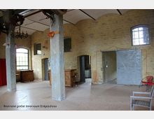 Innenraum einer ehemaligen Brennerei mit gelben Backsteinwänden, großen Bogenfenstern, massiven Betonpfeilern und einer Mischung aus industriellen und rustikalen Elementen. Im Vordergrund stehen Möbel, und an der Decke hängen alte Maschinenteile sowie ein Kronleuchter.