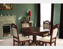 Esszimmer mit einem runden Holztisch und vier gepolsterten Stühlen in klassischem Stil, vor einem grün gestrichenen Kamin mit feinen Ornamenten. Im Hintergrund ein weißer Vorhang vor einem großen Fenster, eine Tischlampe und Wanddekorationen.