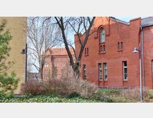 Ziegelgebäude mit hohen Fenstern und Spitzbogen-Elementen, umgeben von kahlen Bäumen, Sträuchern und einem bepflanzten Bereich; im Hintergrund ein weiteres Backsteingebäude mit ähnlicher Architektur. Links im Bild ist eine Mauer aus gelben Ziegeln zu sehen.