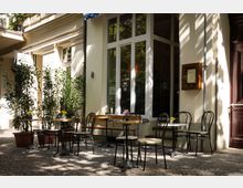 Außenbereich eines Cafés mit mehreren kleinen Tischen und schwarzen Stühlen auf einem gepflasterten Boden. Umgeben von Pflanzen in Töpfen und einem beigen Gebäude mit großen Fenstern, darunter ein Holzrahmen mit einer Speisekarte.