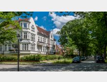 Straßenszene mit einer Reihe von historischen Wohnhäusern mit verzierten Fassaden und roten Dächern. Bäume säumen die gepflasterte Straße, an der Autos geparkt sind, und Straßenschilder zeigen die Namen „Fürstenstraße“ und „Beerenstraße“.