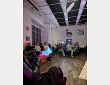 Spacious indoor event room with wood flooring, rows of green chairs, and white walls, featuring large windows on one side. The high ceiling has exposed pipes and acoustic panels, and the back wall displays a mounted antelope and a “Price & Son” sign.