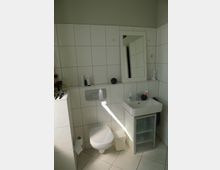Kleines Badezimmer mit weißen Fliesen an den Wänden und auf dem Boden. Links befindet sich eine Toilette mit darüberliegender Spülung, daneben ein kleines Waschbecken mit Spiegel und Ablagefläche, auf der verschiedene Utensilien stehen.