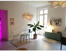 Helles Wohnzimmer mit weißen Wänden, großem Fenster mit Glastür, modernem Kronleuchter und minimalistischer Einrichtung. Der Raum enthält zwei Klappstühle, einen kleinen runden Tisch, eine grüne Kommode, eine Zimmerpflanze in einem Topf und einen hellen Teppich.