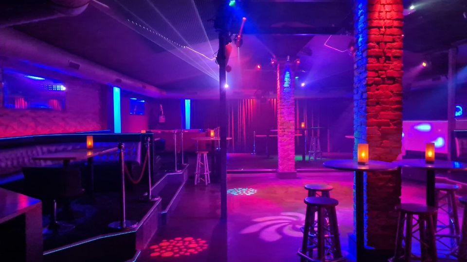 Innenraum eines Nachtclubs mit farbiger LED- und Laserbeleuchtung, Backsteinsäulen und niedriger Decke mit sichtbaren Lüftungsrohren. Hohe Stehtische mit Hockern und kleinen Kerzen sowie gepolsterte Sitznischen säumen eine freie Tanzfläche.