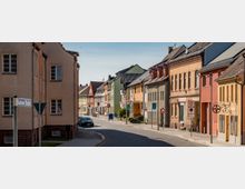 Eine Straße in einer Kleinstadt mit zweistöckigen Wohn- und Geschäftsgebäuden in unterschiedlichen Farben. Die Straße ist gepflastert, gesäumt von geparkten Autos und Straßenschildern, und eine Mischung aus modernisierten und traditionellen Fassaden ist sichtbar.