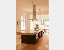 Moderne Küche mit heller hölzerner Fischgrätenbodenstruktur, einer großen Kochinsel mit Betonarbeitsplatte, integriertem Herd und Schränken. Im Hintergrund sind hohe Fenster, ein Esstisch mit roten Stühlen sowie Hängelampen und eine Dunstabzugshaube sichtbar.