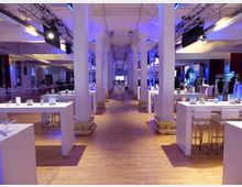 Großer, modern eingerichteter Veranstaltungsraum mit einer zentralen Reihe von weißen Säulen, hohen Decken und einer blauen Beleuchtung. Der Raum ist mit weißen Tischen und Barhockern ausgestattet, und im Hintergrund sind Messe- oder Ausstellungselemente zu sehen.