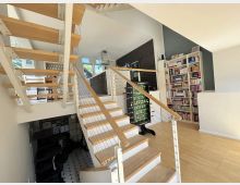 Offener Wohnbereich mit zentraler Holztreppe und Geländer aus Seilen, daneben eine moderne Küche mit dunklen Schränken. Rechts steht ein großes, weißes Bücherregal; der Raum hat helle Holzfußböden und große Fenster.