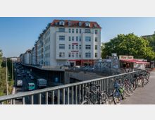 Straßenszene mit einem mehrstöckigen Wohn- und Geschäftshaus, das ein Hotel beherbergt, und einem angrenzenden Imbissstand mit der Aufschrift „Enjoy Kebap“. Im Vordergrund stehen Fahrräder an einem Geländer, darunter verläuft eine befahrene Straße. Im Hintergrund sind Bäume und weitere Gebäude sichtbar.