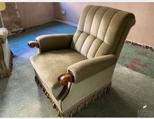 Innenraum eines älteren, teils abgenutzten Zimmers mit beigefarbenem Polstersessel mit Holzarmlehnen und Fransenbesatz. Der Boden ist mit grauem Teppich und sichtbaren blau-grünlichen, abgenutzten Flächen bedeckt; an den Wänden sind Gebrauchsspuren und eine Steckdose zu sehen.