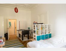 Helles Kinderzimmer mit hohen Decken und Stuckverzierungen, eingerichtet mit einem Bett, einem Bücherregal, einem Tischkicker und einer bunten Teppichmatte. Durch eine offene Tür ist ein angrenzender Flur sichtbar.