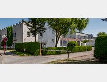 Ein modernes, einstöckiges Gebäude mit wellenförmiger Dachkante auf der linken Seite und einer Glasfassade auf der rechten Seite, umgeben von gepflegten Hecken und Bäumen. Im Vordergrund befindet sich eine Schranke, die eine gepflasterte Zufahrt begrenzt.