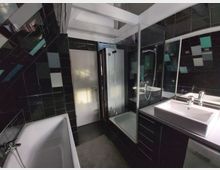 Modernes Badezimmer mit dunklen, glänzenden Fliesen und weißen Elementen. Es enthält eine Badewanne, eine Dusche mit Glastür, ein Waschbecken mit Spiegel sowie ein weißes Türmodell im Hintergrund.