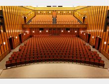 Großer Zuschauerraum eines Theaters oder Auditoriums mit zahlreichen gepolsterten, orangefarbenen Sitzreihen, zwei Ebenen und holzverkleideten Wänden. Das Bild zeigt den Blick von der Bühne in den Saal, der von warmem Licht beleuchtet wird.