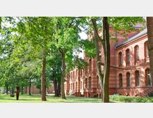 Backsteinbau im neogotischen Stil mit großen Fenstern und Details aus helleren Ziegeln, umgeben von einem parkähnlichen Gelände mit Rasen, Bäumen und einzelnen modernen Skulpturen.