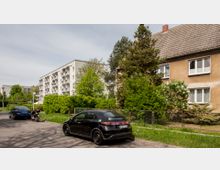 Straßenszene in einem Wohngebiet mit geparkten Autos und einem Motorrad. Rechts ein älteres Wohnhaus mit brauner Fassade und Satteldach, links ein moderneres Mehrfamilienhaus mit Balkonen und heller Fassade, umgeben von Bäumen und Hecken.