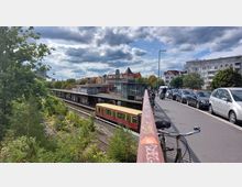 S-Bahnhof in städtischer Umgebung mit einem gelben und roten Zug auf den Gleisen. Im Vordergrund eine Brücke mit geparkten Fahrrädern und Autos, umgeben von Bäumen und Wohnhäusern im Hintergrund.
