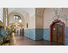 Langer Korridor eines historischen Gebäudes mit dekorativen Elementen, darunter verzierte Holz-Doppeltüren mit einem Relief und türkisfarbene Wandfliesen. Die Decke ist gewölbt, große Fenster auf der linken Seite lassen Tageslicht einfallen, und Sitzbänke stehen entlang der Wand.