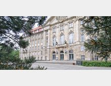 Historisches Gebäude mit symmetrischer Fassade, großen Rundbogenfenstern und verzierter Architektur, flankiert von Säulen und Skulpturen. Im Vordergrund sind Zweige eines Nadelbaums zu sehen, die teilweise den Blick auf das Gebäude einrahmen.