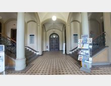 Eingangsbereich eines historischen Gebäudes mit verzierten Säulen, Rundbogenarchitektur und einem gefliesten Boden. Zu beiden Seiten führen Treppen mit kunstvollen Geländern hoch, und in der Mitte befindet sich eine Holztür; im Vordergrund ein Prospektständer mit Informationsmaterial.