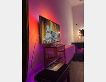 Interior of a modern living/dining room with a wall-mounted flat-screen TV showing a fireplace, lit by red and purple ambient lighting. Below the TV is a small wooden cart with shelves, a radiator along the wall, and a dining table with pendant lights in the background.