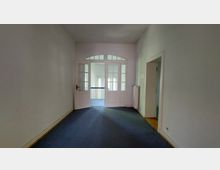 Flur eines Gebäudes mit blauem Teppichboden, weißen Wänden und einer Doppeltür aus Glas mit Sprossen. Hinter der Tür ist ein weiterer Bereich mit großen Fenstern und Geländer erkennbar.