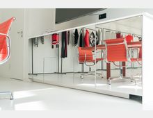 Ein heller Raum mit weißem Boden und einem großen Spiegel, der einen Teil eines Konferenz- oder Ankleidezimmers widerspiegelt. Sichtbar sind rot-orange Drehstühle an einem Tisch sowie Kleidungsstücke an einer Kleiderstange im Hintergrund.