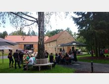 Ein Hof mit mehreren Gebäuden aus hellem Backstein und roten Ziegeldächern, umgeben von Bäumen und Wiesen. Im Vordergrund sitzen Menschen an Tischen unter Sonnenschirmen, mit einem Baum, der von einer Bank umgeben ist.