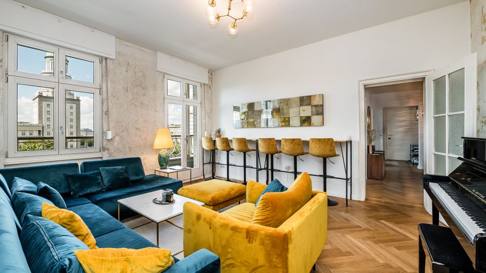 Helles Wohnzimmer mit großen Fenstern, freiliegender Putzwand und Fischgrätparkett, eingerichtet mit einer blauen Eckcouch, gelbem Sessel und Couchtisch. An der Wand steht eine schmale Bar mit hohen Stühlen, rechts neben einer Glastür ist ein Klavier zu sehen.