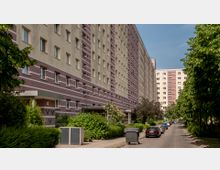 Mehrstöckiges Wohngebäude im Stil eines Plattenbaus mit cremefarbenen und rötlichen Fassadenstreifen, flankiert von Bäumen und Büschen. Im Vordergrund führt eine schmale Straße mit parkenden Autos und einer Mülltonne entlang.