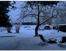 Verschneiter Garten mit großem, schneebedecktem Baum im Vordergrund und Blick auf ein weißes Feld und einen dunklen Waldrand in der Ferne. Rechts stehen zwei Stühle und ein niedriger Tisch im Schnee, daneben eine Schaukel und ein kleines Gartenhäuschen.