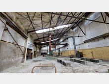 Eine verlassene Industriehalle mit hohen Wänden, Stahlträgern an der Decke und oberseitigen Lichtfenstern. Die Räume sind teils mit gelben Fliesen verkleidet, und es befinden sich alte Rohre, Maschinenreste und Betonfundamente auf dem Boden.