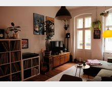 Gemütliches Wohnzimmer mit Holzfußboden, großem Fenster und Glastür, einem TV auf niedrigem Sideboard sowie einem Plattenschrank mit Schallplatten. An den Wänden hängen Bilder, von der Decke hängen Pflanzen und mehrere Pendelleuchten über Sofa und Couchtisch.