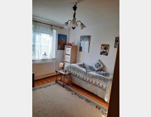 Ein kleines Schlafzimmer mit Einzelbett, das mit einer blauen Patchwork-Tagesdecke und einem passenden Kissen dekoriert ist. Neben dem Bett stehen ein kleiner Beistelltisch und ein schmaler Holzschrank, das Zimmer hat helle Wände, ein großes Fenster mit durchscheinenden Gardinen und ist mit einem Teppich auf Holzboden ausgestattet.