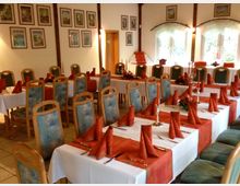 Ein festlich gedeckter Veranstaltungsraum mit mehreren Tischen, die mit weißen Tischdecken, orangefarbenen Tischläufern und passend gefalteten Stoffservietten dekoriert sind. Die Holzstühle mit gepolsterten, gemusterten Sitzflächen stehen in Reihen, und an den Wänden hängen gerahmte Bilder unterhalb bogenförmiger Fenster mit weißen Gardinen.