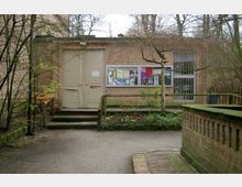 Ein einstöckiges Gebäude aus Backstein mit einem beigen Doppeltor und einem Infokasten an der Wand, der mit Plakaten und Aushängen gefüllt ist. Im Vordergrund befinden sich ein gepflasterter Weg, kleine Beete mit Blumen und Sträucher sowie ein Geländer.