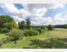 Weitläufige Grünfläche mit vereinzelten Bäumen, Sträuchern und einem Holzzaun im Hintergrund. Der Himmel ist teils bewölkt, und die Umgebung vermittelt den Eindruck einer ländlichen Parklandschaft oder eines Gartens.
