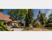 Ein ruhiger Dorfplatz mit einer kleinen Kapelle rechts im Hintergrund, gekennzeichnet durch einen Kirchturm mit einer Wetterfahne, umgeben von Bäumen. Links befindet sich ein Wohnhaus mit einem roten Ziegeldach und bewachsenem Eingang, sowie ein gepflasterter Weg, der zwischen den Gebäuden verläuft.