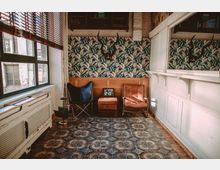 Wohnraum im Vintage-Stil mit gemustertem Fliesenboden, einer Tapete mit Blattmotiv, zwei Sesseln (einer aus Leder, einer mit schwarzer Stofflehne) und einer weißen Holzverkleidung an der Wand. Ein Geweih hängt an der Rückwand, und durch ein großes Fenster links fällt Tageslicht ein.