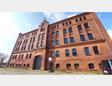 Backstein-Gebäude mit mehreren Stockwerken und großen Fenstern im industriellen Baustil. Vor dem Gebäude befinden sich ein gepflasterter Platz, ein Basketballkorb und ein parkendes Fahrzeug.