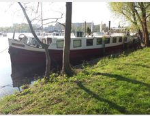 Hausboot mit weißem Aufbau und rotem Rumpf, am Ufer eines Gewässers vertäut. Im Vordergrund sind grüne Wiesen und Bäume, während im Hintergrund moderne Gebäude und weitere Boote sichtbar sind.