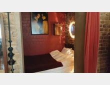 Kleines Schlafzimmer mit roten Backstein- und Fliesenwänden, dunkelroten Vorhängen und einem Doppelbett. Ein gerahmtes Gemälde, ein verzierter Spiegel sowie ein kleines Holzschränkchen dekorieren den Raum.