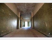 Langer, schmaler Innenkorridor mit abgenutzten grün-beigen Wänden, Betonboden mit eingelassenen Rinnen und einer niedrigen Decke. Mehrere Stützpfeiler gliedern den Raum, am Ende führt eine offene Tür in einen hellen Außenbereich.