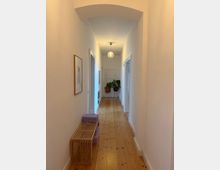 Long, narrow residential hallway with white walls, a light wood plank floor, and a shallow arched ceiling near the foreground. A globe pendant light hangs overhead, with doorways along both sides, a wooden slatted bench and framed wall art on the left, and potted plants near a closed door at the far end.