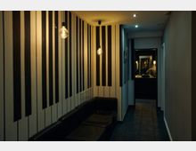 Ein schmaler Gang mit Wänden, die mit schwarzen und weißen vertikalen Streifen dekoriert sind. Links stehen schwarze Sitzbänke, an der Decke hängen freiliegende Glühbirnen, und am Ende des Gangs ist ein heller Bereich mit Spiegel und dekorativen Elementen sichtbar.